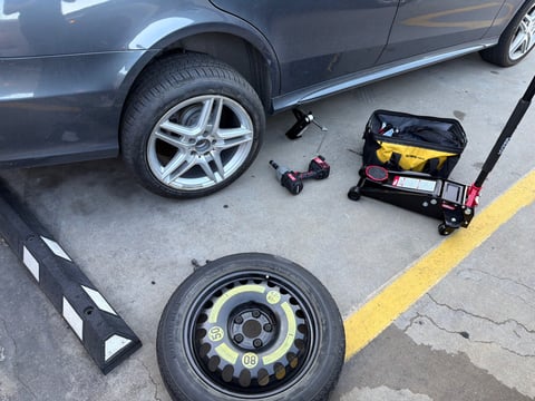 Gray Mercedes-Benz with AMG-style alloy wheel removed and set on a parking block, OEM donut spare staged on the ground, Hyper Tough 3-ton floor jack and cordless impact in a clean Austin TX parking lot — All Hours Roadside mobile tire change