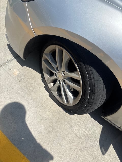 Silver Chevrolet sedan with a Firestone Firehawk tire destroyed by a sidewall blowout — chunks of rubber missing along the outer sidewall, mounted on OEM alloy wheel — All Hours Roadside mobile tire replacement in Austin, TX