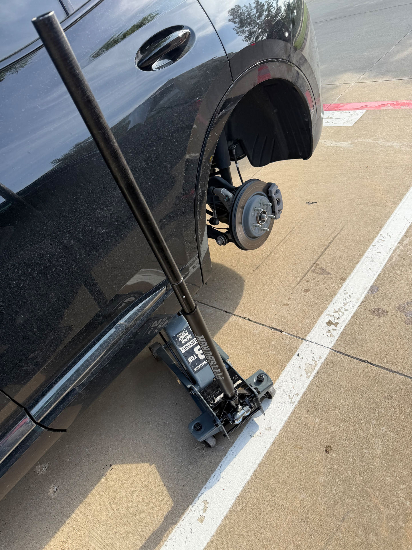 Black SUV raised on a red Pittsburgh three-ton floor jack with the rear wheel removed during a parking-lot spare tire change in Grand Prairie, Texas