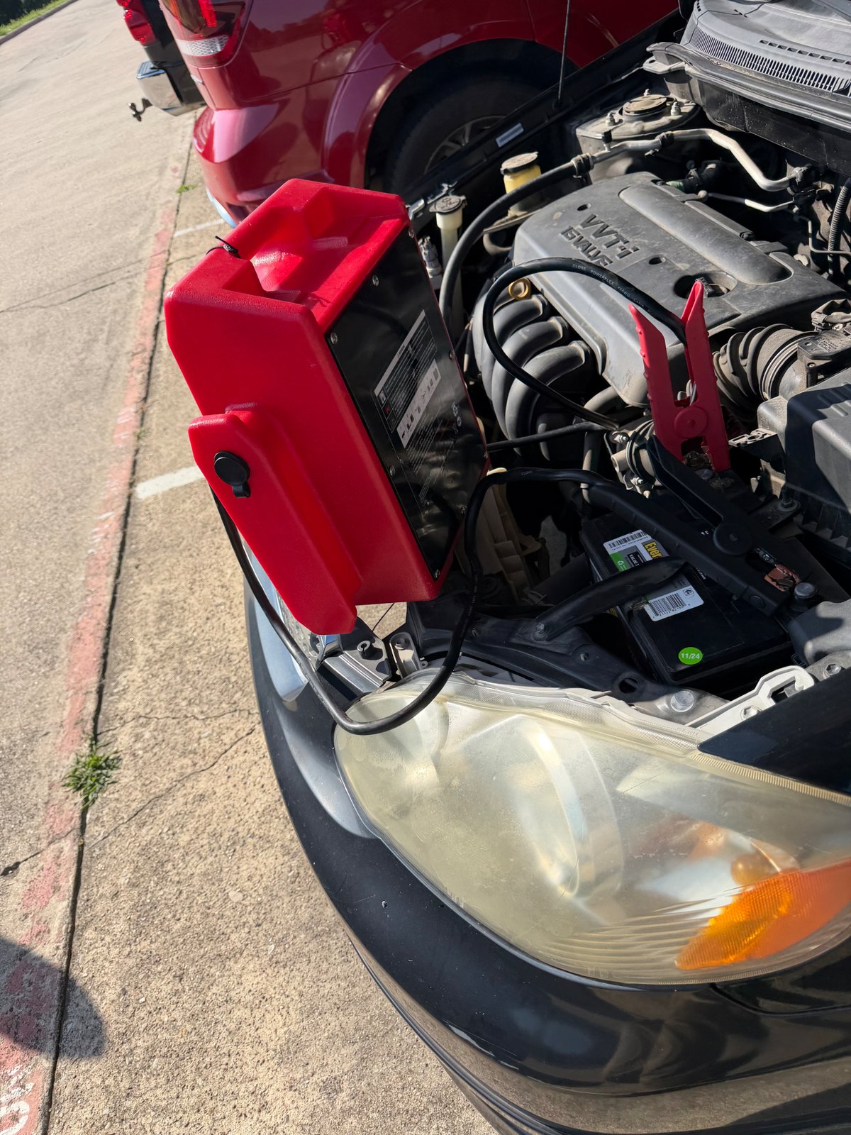 Red commercial jump pack clamped to a Hyundai 12V battery on S. Cooper Street in Arlington TX with a red Ford F-150 parked behind — All Hours Roadside 24/7 mobile jump start