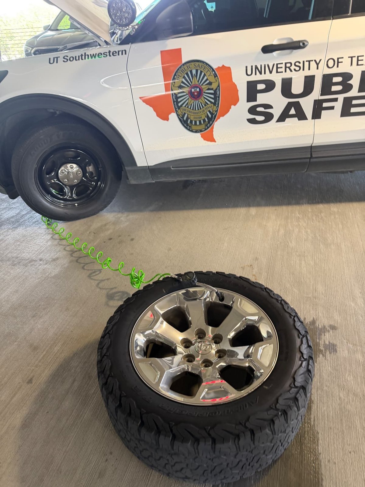 Full-size Ram truck wheel with a chrome alloy rim and all-terrain tire laid on the concrete of a Dallas campus parking deck, green coiled air hose and inflator chuck attached to the valve stem, with a white university public safety Ford Explorer patrol SUV on a black steel spare wheel in the background during a mobile fleet tire change by All Hours Roadside