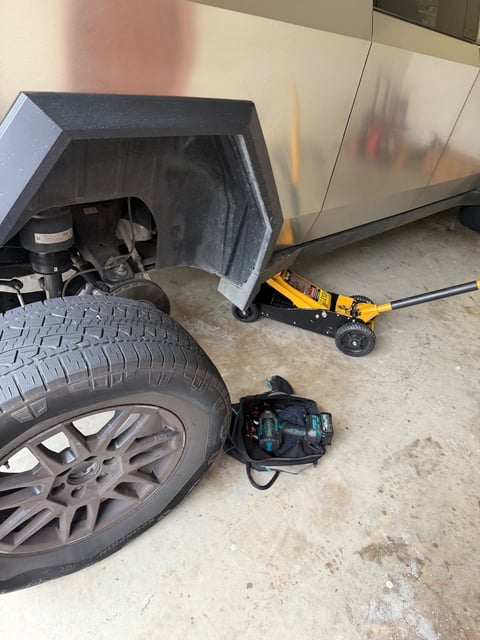 Tesla Cybertruck raised with a yellow floor jack in a garage, wheel removed and staged beside a tool bag