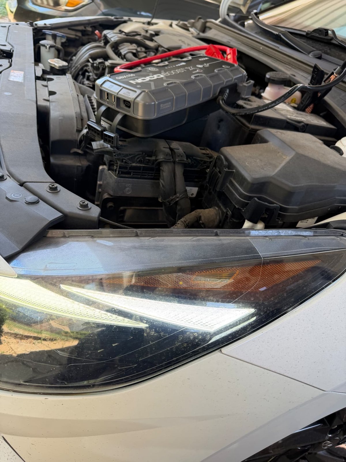NOCO Boost X GBX155 lithium jump pack resting on the engine cover of a silver sedan with the hood raised and LED headlight lit during a mobile jump start in Lewisville TX by All Hours Roadside
