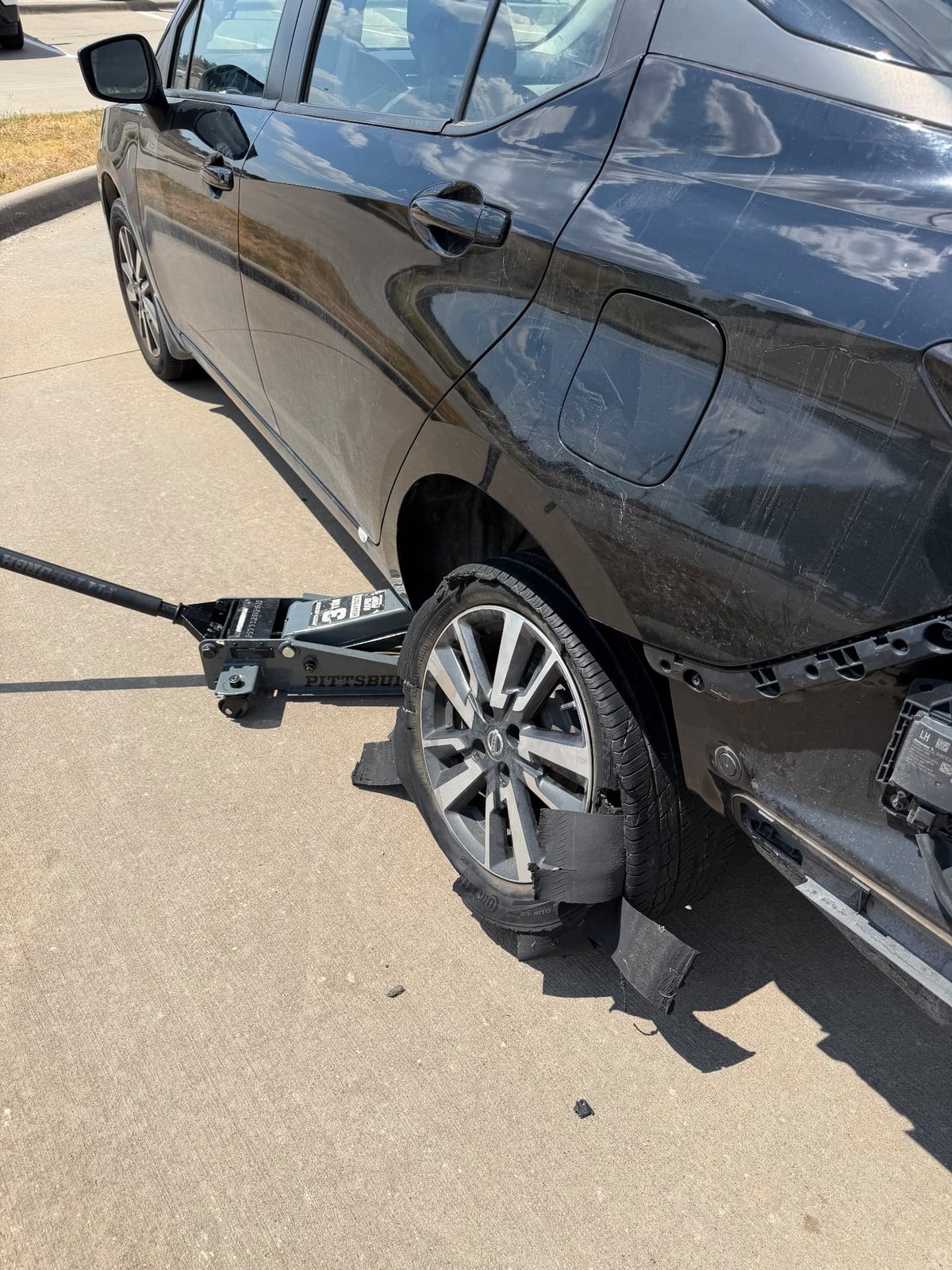 Black Nissan sedan lifted on a Pittsburgh floor jack in a Dallas TX parking lot with a completely shredded rear tire, tread chunks scattered on the concrete, during an on-site mobile tire change by All Hours Roadside