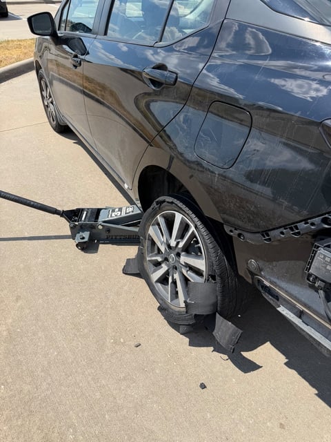 Black Nissan sedan lifted on a Pittsburgh floor jack in a Dallas TX parking lot with a completely shredded rear tire, tread chunks scattered on the concrete, during an on-site mobile tire change by All Hours Roadside