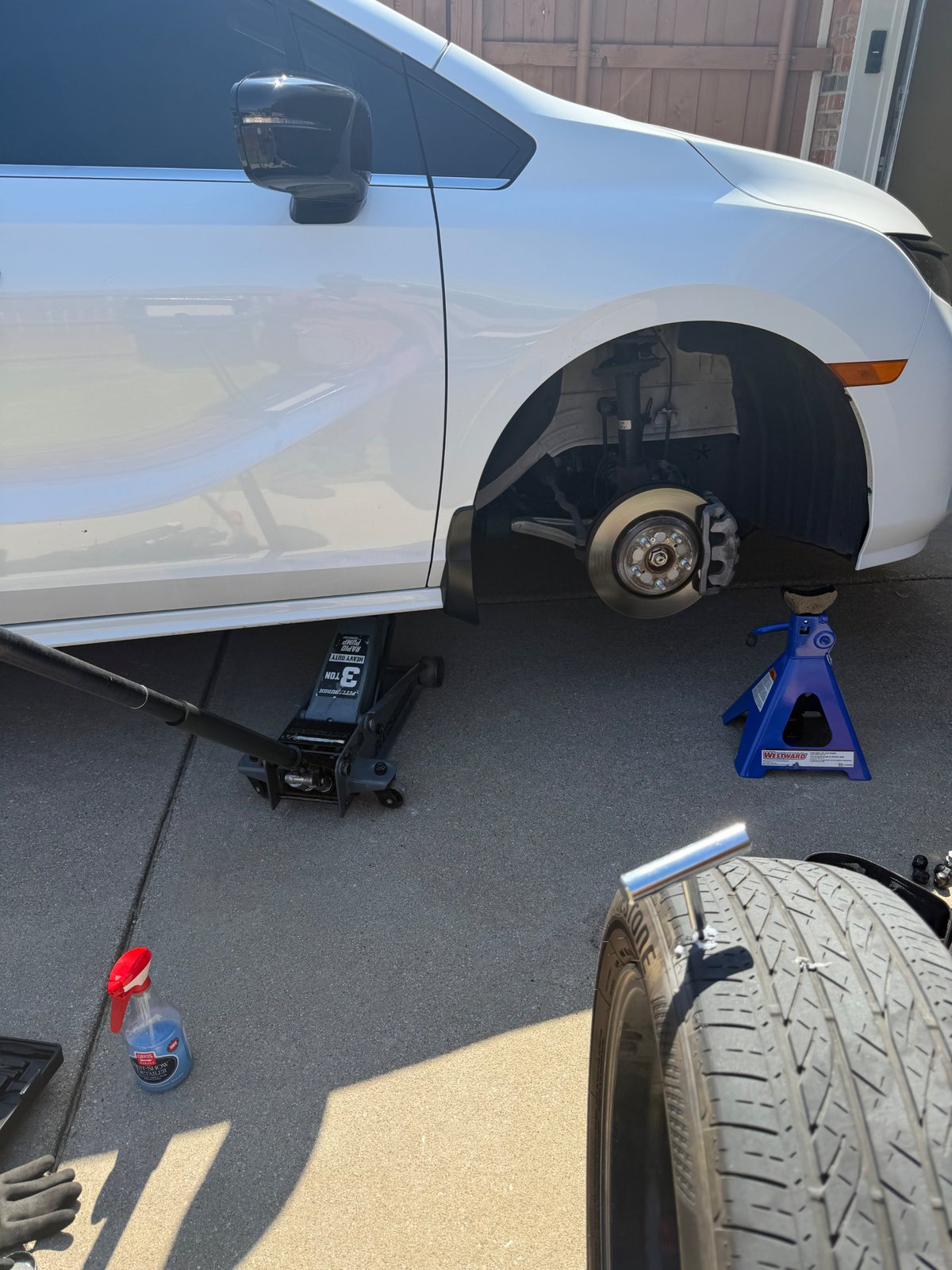 All Hours Roadside technician changing a front tire on a white Chrysler Pacifica in an Allen, TX driveway — 3-ton Pittsburgh floor jack and Westward jack stand supporting the vehicle, Bridgestone tire staged for install.