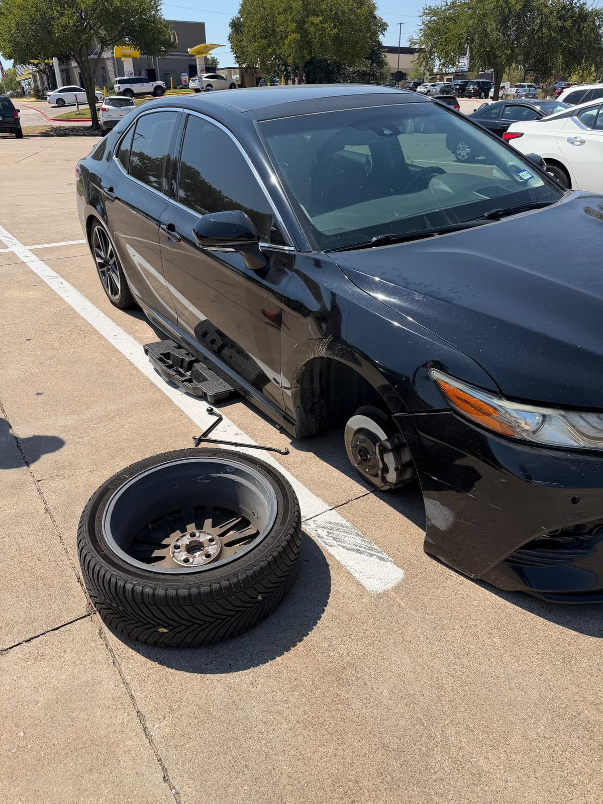 Black Toyota Camry raised on a floor jack in a Dallas retail parking lot with the front alloy wheel removed and a cross-wrench on the pavement during a mobile tire change by All Hours Roadside