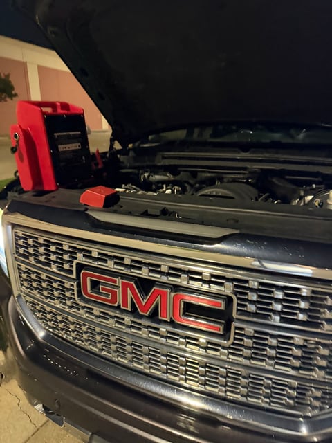 Red heavy-duty portable jump pack set on the radiator support of a black GMC Sierra Denali HD with the hood raised at night in Irving TX — All Hours Roadside mobile jump start
