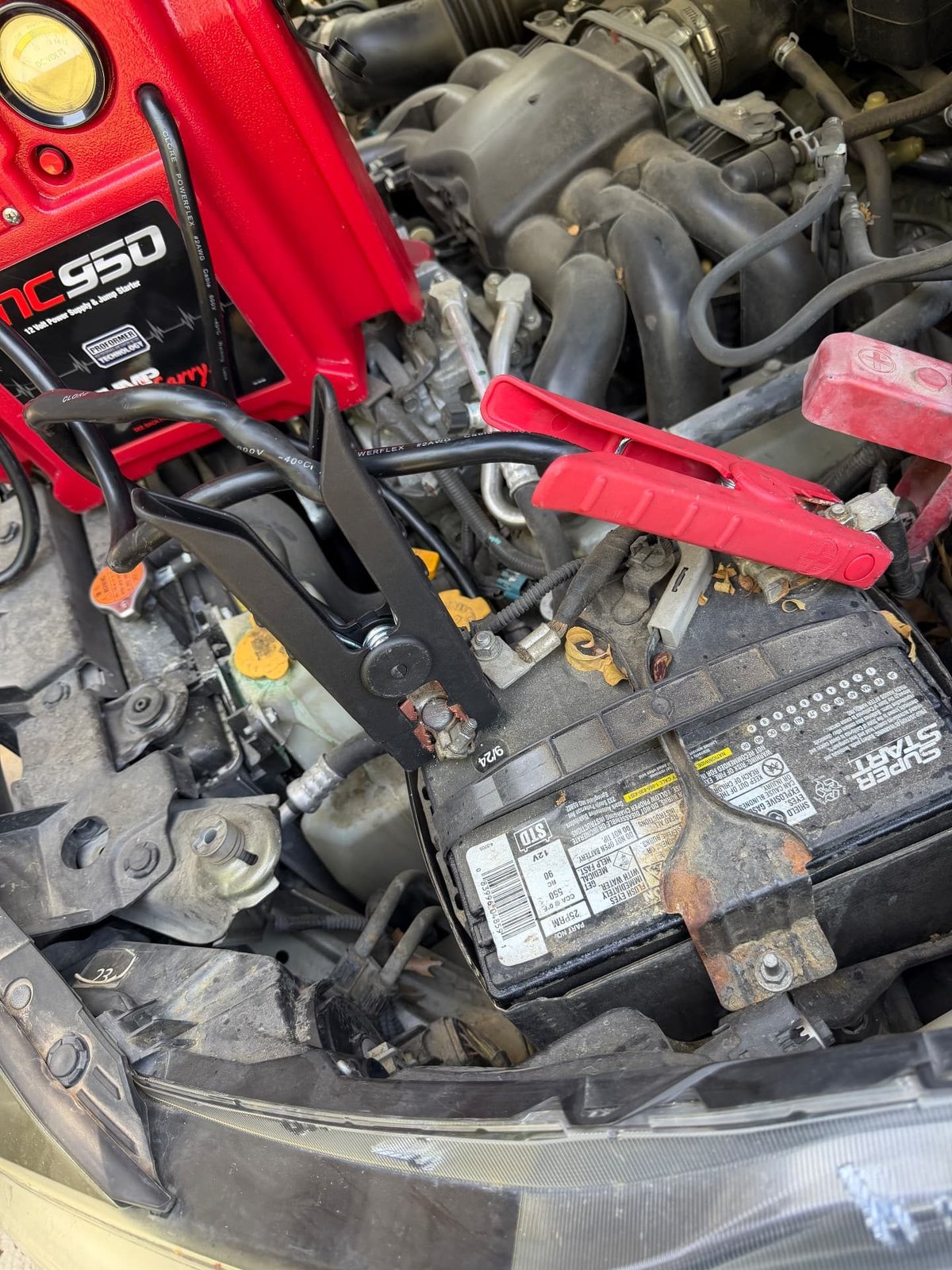 Red Clore JNC950 commercial jump pack clamped to a corroded Super Start 12V battery with a 9/24 date code and white terminal buildup under the hood during a mobile battery service call in Dallas Fort Worth by All Hours Roadside