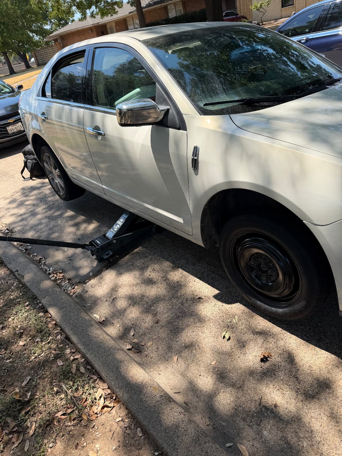 White Lincoln MKZ raised on a black floor jack on a residential street in Irving TX with the temporary donut spare mounted on the rear hub during a mobile tire change by All Hours Roadside