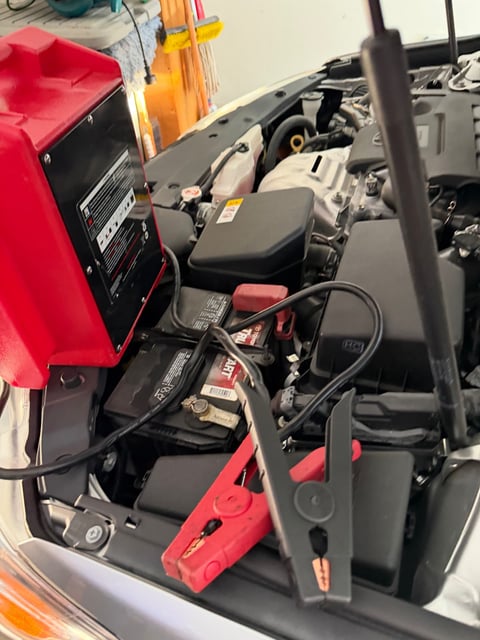 Red commercial jump pack clamped to a Toyota TruStart 12V battery in the engine bay of a silver Toyota with the hood raised in McKinney TX — All Hours Roadside mobile jump start