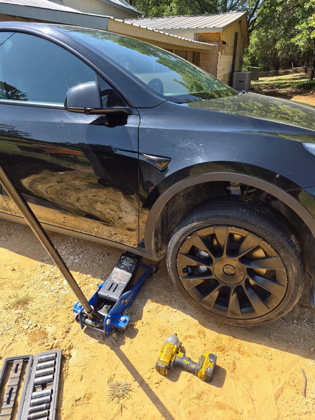 Black Tesla Model Y lifted at the front-right pinch-weld jack point with a blue low-profile floor jack on a gravel driveway in DFW Black Tesla Model Y lifted at the front-right pinch-weld jack point with a blue low-profile floor jack on a gravel driveway in DFW