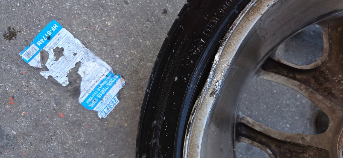 Heavy curb rash and gouging on a multi-spoke aluminum alloy wheel next to a torn ST205/75R15 tire label — inspected by All Hours Roadside in the Dallas–Fort Worth metro