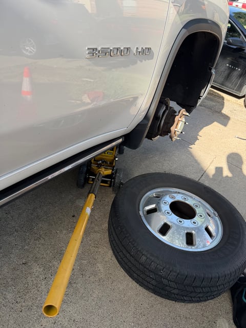 Silver Chevrolet Silverado 3500 HD dually pickup truck lifted on a heavy-duty CAT bottle jack with the rear wheel removed and a Firestone Transforce HT spare staged on the ground during a mobile heavy-duty tire change by All Hours Roadside in Dallas, TX